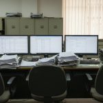 An empty back-office workstation with three monitors showing spreadsheets and database records, surrounded by thick stacks of paper and rows of olive-green filing cabinets.
