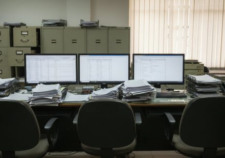 An empty back-office workstation with three monitors showing spreadsheets and database records, surrounded by thick stacks of paper and rows of olive-green filing cabinets.