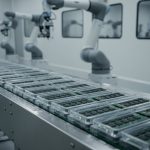Circuit boards in clear plastic trays moving along a conveyor in a cleanroom, with white robotic arms positioned along the line.