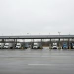 A row of transport trucks parked under a long canopy at a quiet border inspection plaza on a wet, overcast day.