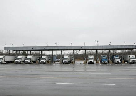 A row of transport trucks parked under a long canopy at a quiet border inspection plaza on a wet, overcast day.