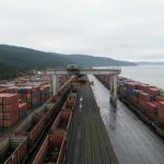 Aerial view of a rain-soaked container terminal beside a forested coastal inlet, with stacked shipping containers and freight rail cars flanking a gantry crane over the tracks.