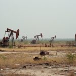 Several rusting oil pumpjacks and a corroded storage tank standing in a dry, scrubby field littered with abandoned pipe and debris under a pale hazy sky.