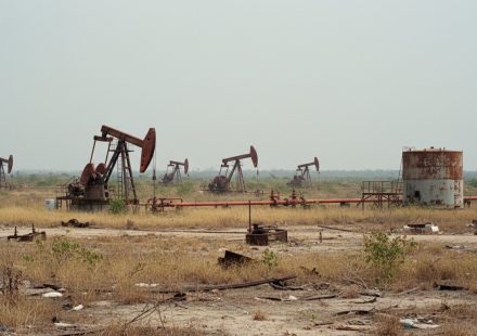 Several rusting oil pumpjacks and a corroded storage tank standing in a dry, scrubby field littered with abandoned pipe and debris under a pale hazy sky.