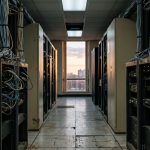 A dimly lit corridor of aging server racks with bundled cabling, one dead ceiling light panel, and warm sunset light through a window at the far end.