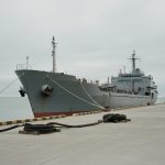 A grey naval support ship moored alongside a bare concrete pier under a flat overcast sky, mooring lines running to a bollard and coiled fuel hoses lying on the empty quay, with no crew in sight.