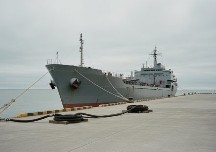 A grey naval support ship moored alongside a bare concrete pier under a flat overcast sky, mooring lines running to a bollard and coiled fuel hoses lying on the empty quay, with no crew in sight.